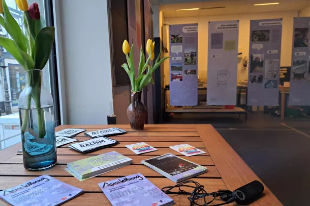 Auf einem Tisch stehen Tulpen und es liegen Flyer und Postkarten aus. Im Hintergrund hängt die Ausstellung 