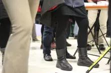 Füße vieler Menschen stehen auf dem Boden der Galerie für die Ausstellungseröffnung. Im Vordergrund ist eine Person in schwarzen Winterstiefeln.