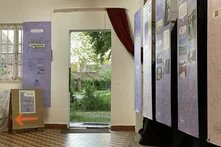 Ausstellungsbanner an Stahlseilen aufgehängt; durch eine offene Tür blickt man raus ins Grüne