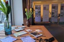 Auf einem Tisch stehen Tulpen und es liegen Flyer und Postkarten aus. Im Hintergrund hängt die Ausstellung "zusammentun – solidarisch gegen rechts an Stahlseilen"