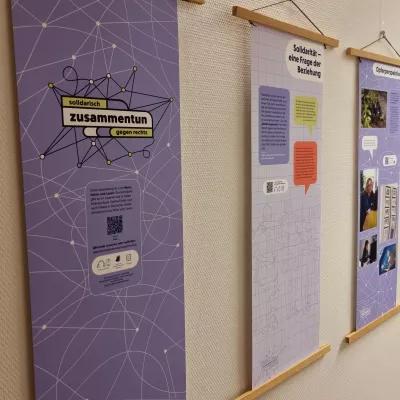 Die Ausstellung "zusammentun" hängt mit Posterleisten aus Holz befestigt an einer Wand.