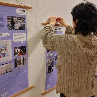 Eine Person mit Strickpullover hängt ein Plakat auf, das in einer Posterleiste klemmt. Daneben hängt bereits ein Plakat.